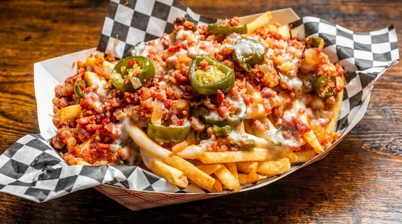 Loaded fries with melted cheese, bacon bits, and sliced jalapeños in a checkered paper tray.